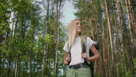 Handheld-shot-of-thoughtful-serious-female-hiker-with-backpack-walking-amidst-trees-in-forest.-A-young-woman-with-a-backpack-walks-through-the-forest-on-a-summer-day.